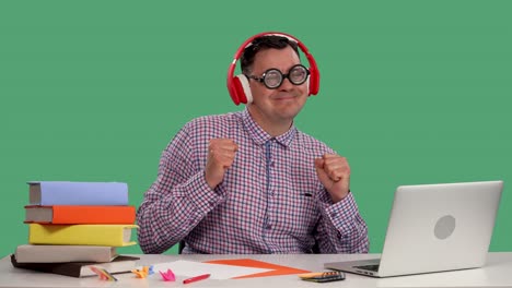 un hombre disfruta de la música en sus grandes auriculares inalámbricos rojos y baila mientras está sentado en una mesa frente a una computadora portátil abierta. retrato de un hombre en el estudio en una pantalla verde. cámara lenta.