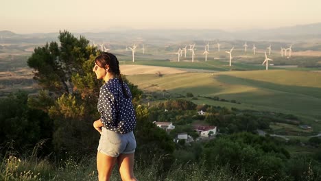 Slowmotion-tracking-shot-of-a-young-woman-walking-up-a-hill-with-windmills-spinning