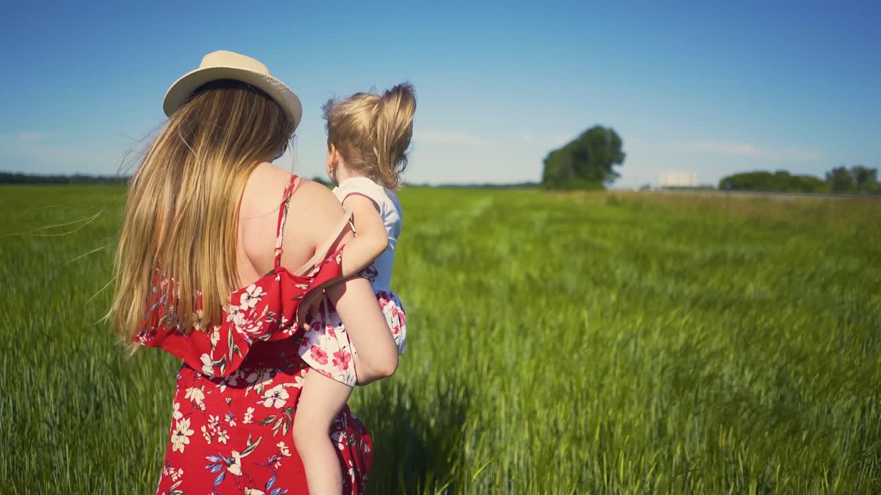 vista de fondo madre en sombrero de verano está caminando a través de un campo sin fin sosteniendo a su hija en sus brazos