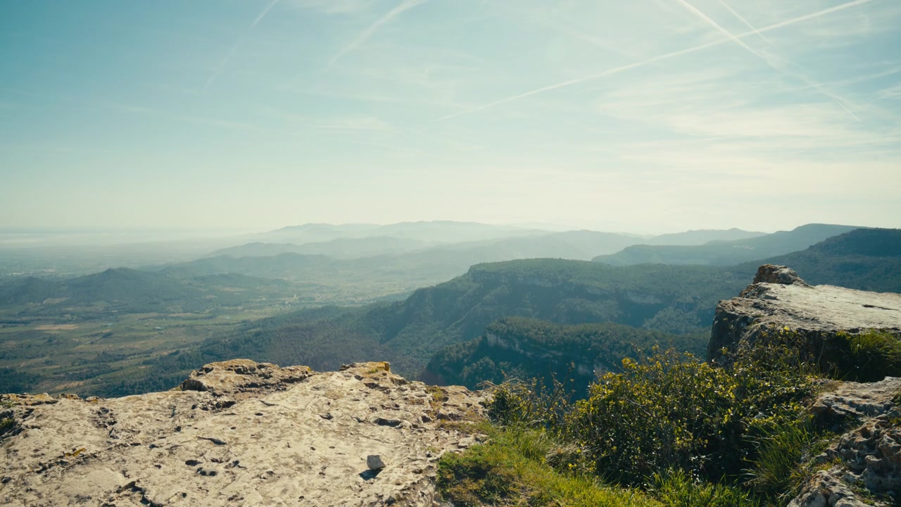 Una sporgenza rocciosa si erge sopra un vasto paesaggio montano, con ripidi strapiombi, creste stratificate e valli distanti che sfumano in una leggera foschia atmosferica in Catalogna, Spagna