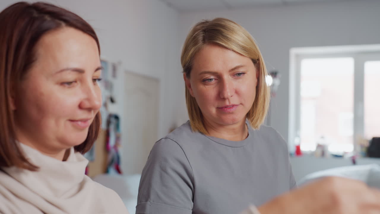 Smiling seamstress showing textile pattern to colleague during cheerful discussion in bright workshop with mood boards and fabrics visible in background, both enjoying creative conversation