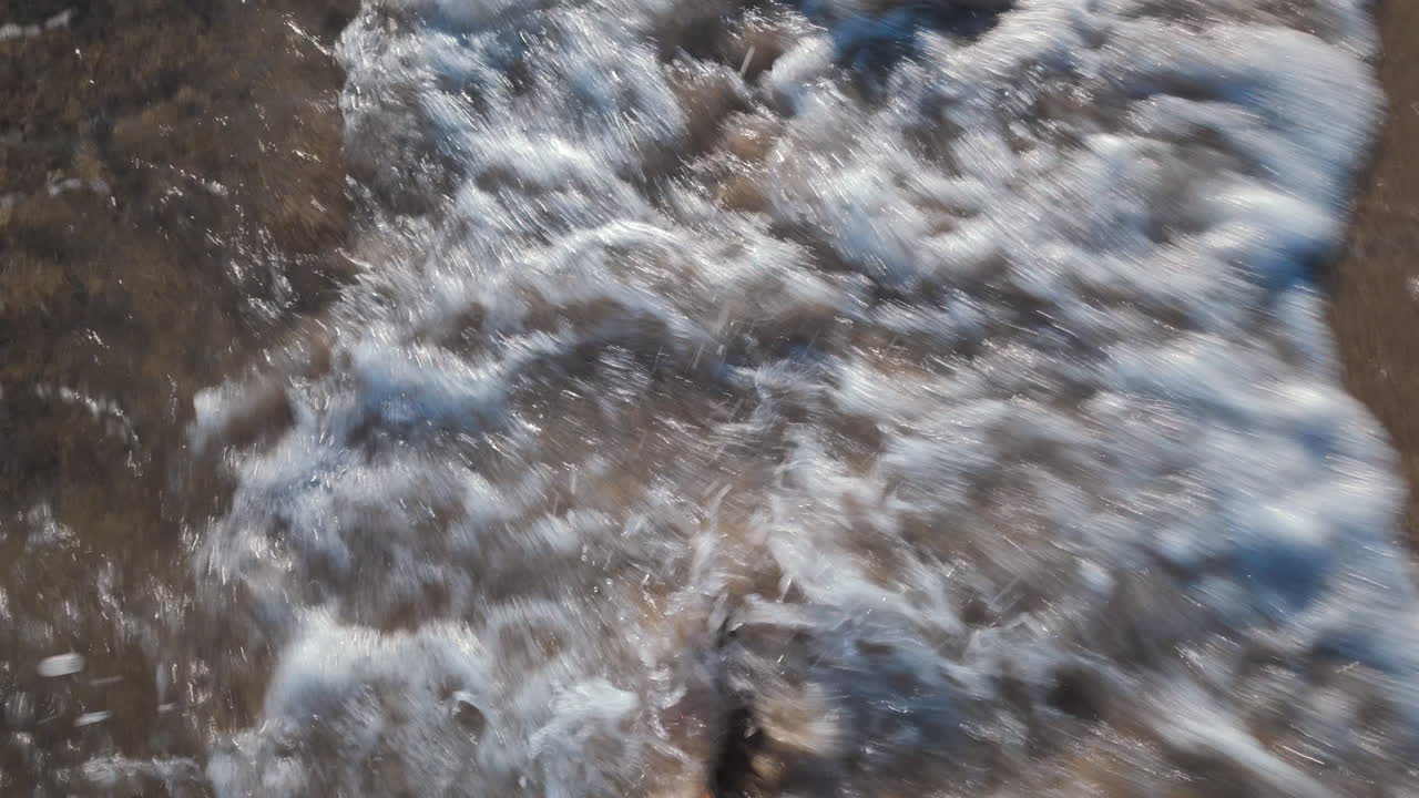 Close up of a man's bare foot touching sea foam while walking on the sandy beach
