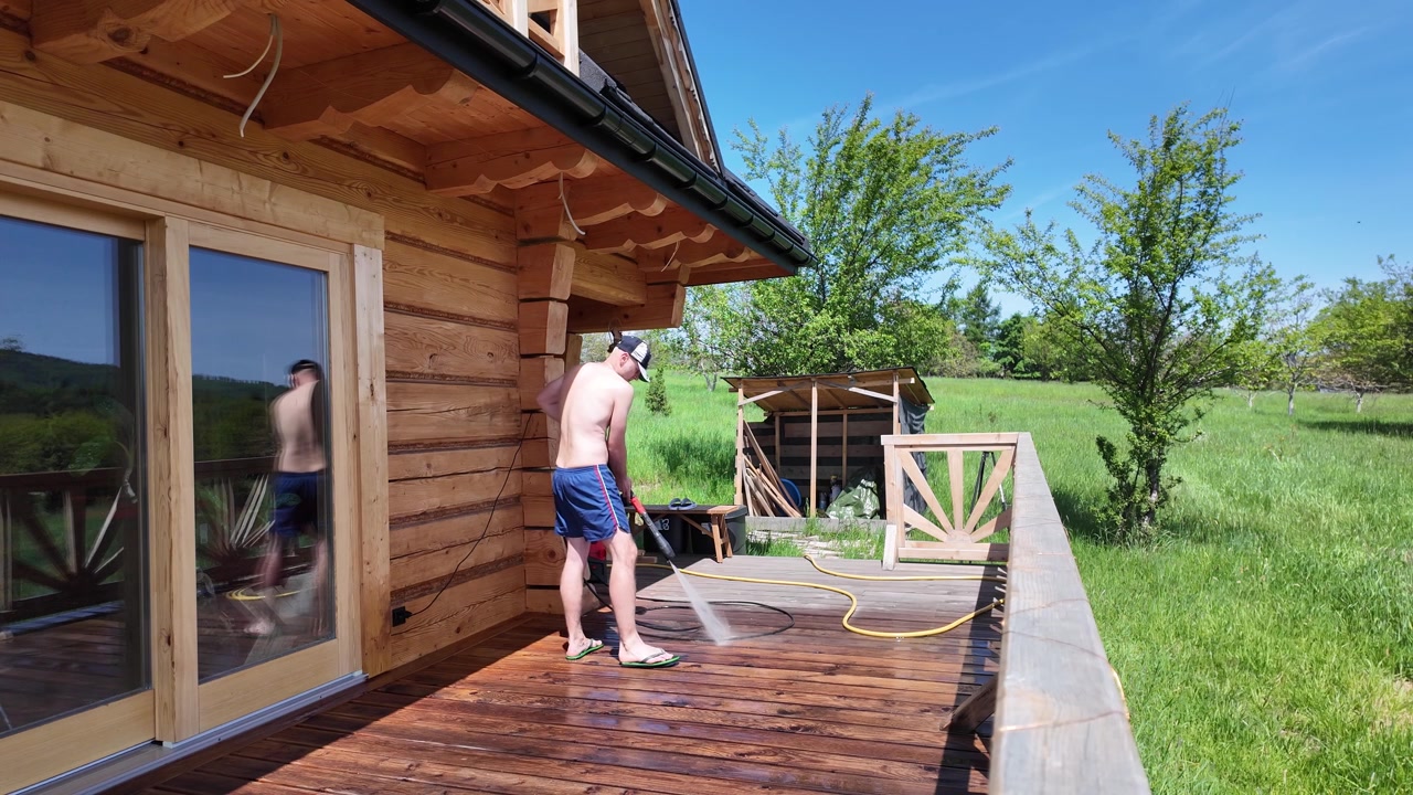 Shirtless worker in blue shorts cleans a wooden terrace with a pressure washer, with green grass, forest, blue sky and mountain peaks visible in the background.