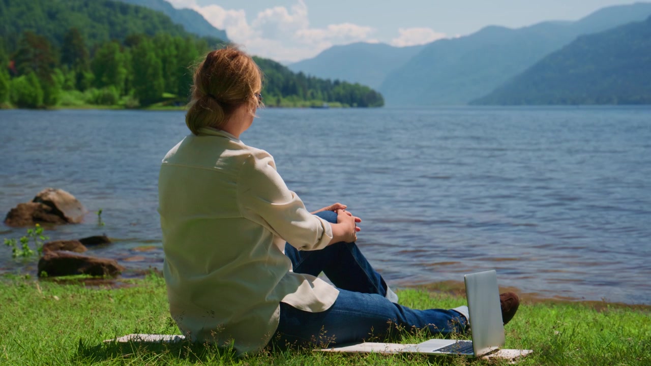 Eine Frau sitzt an ruhigem Wasser, ihr Laptop ist neben ihr geöffnet. Sie wirkt entspannt und konzentriert auf ihre Arbeit oder Gedanken. Berge erheben sich hinter ihr, Bäume säumen das Ufer. Eine friedliche Umgebung, ideal für Remote-Arbeit.