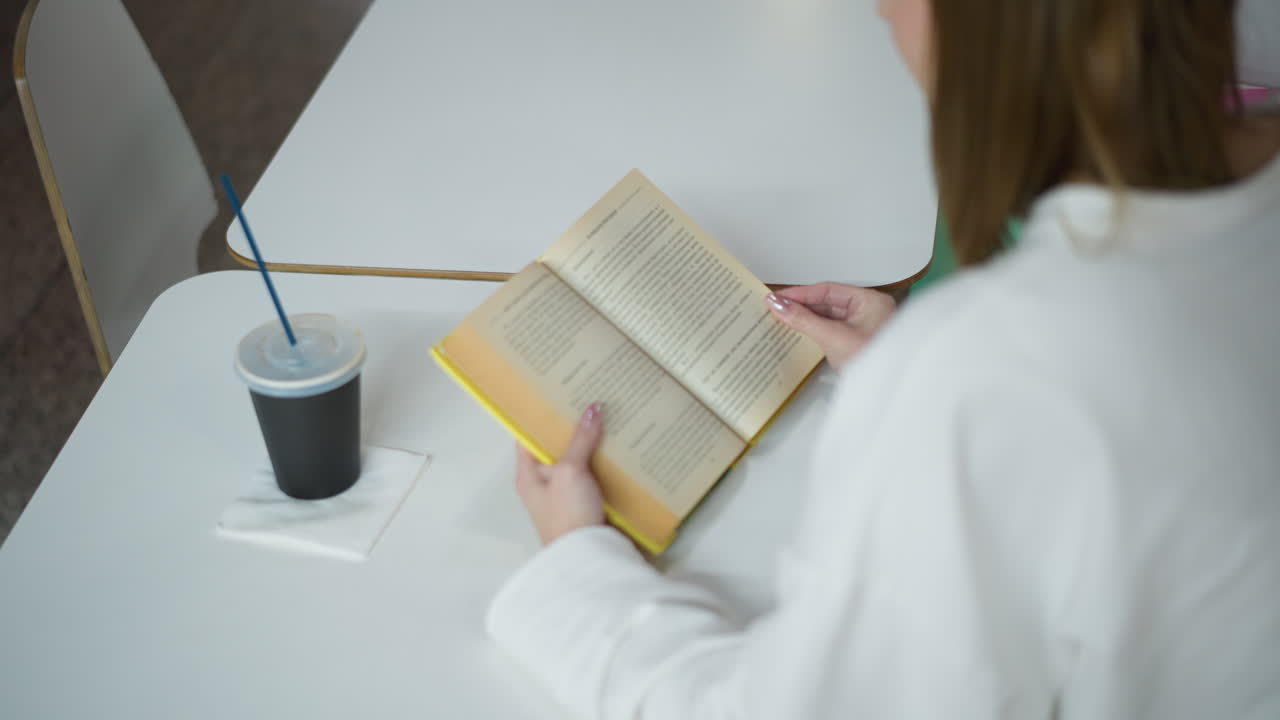 vista aérea de una mujer joven leyendo un libro abierto en una mesa en un centro comercial moderno, con una taza de bebida fría a un lado, entorno relajante con colores brillantes y una estética limpia y minimalista