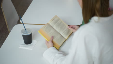 overhead view of young woman reading open book at a table in a modern mall, with a cold drink cup on the side, relaxing environment featuring bright colors and a clean, minimalistic aesthetic