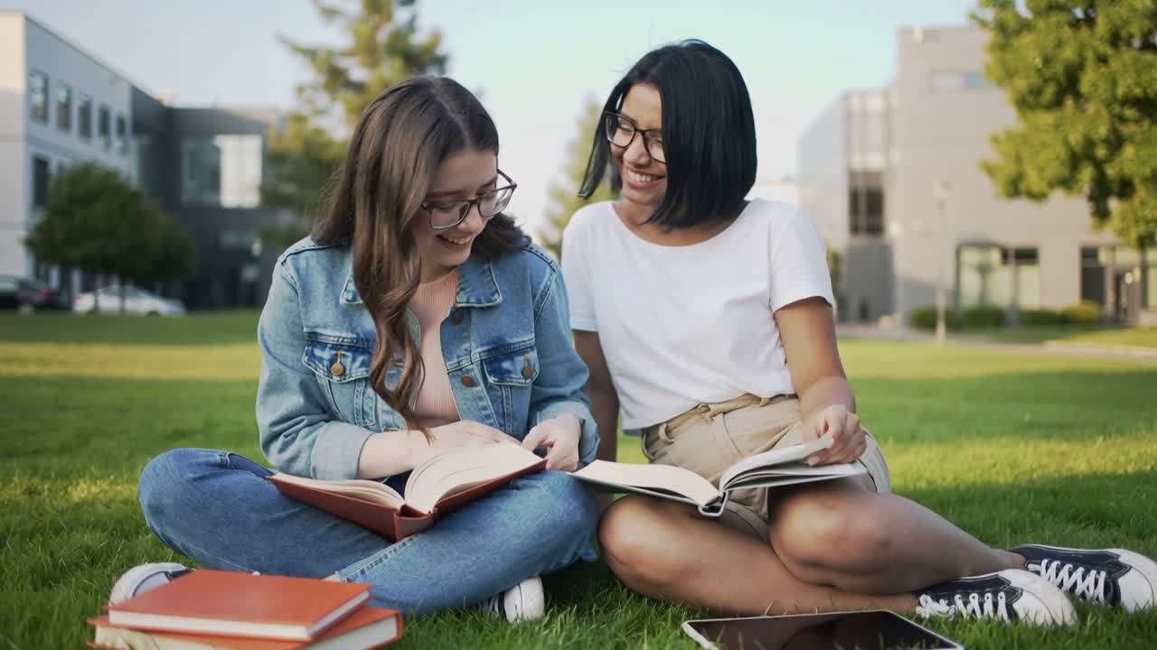 Dos estudiantes sentados en el césped, mujer señalando una página iniciando el estudio con un libro de texto y una tablet.