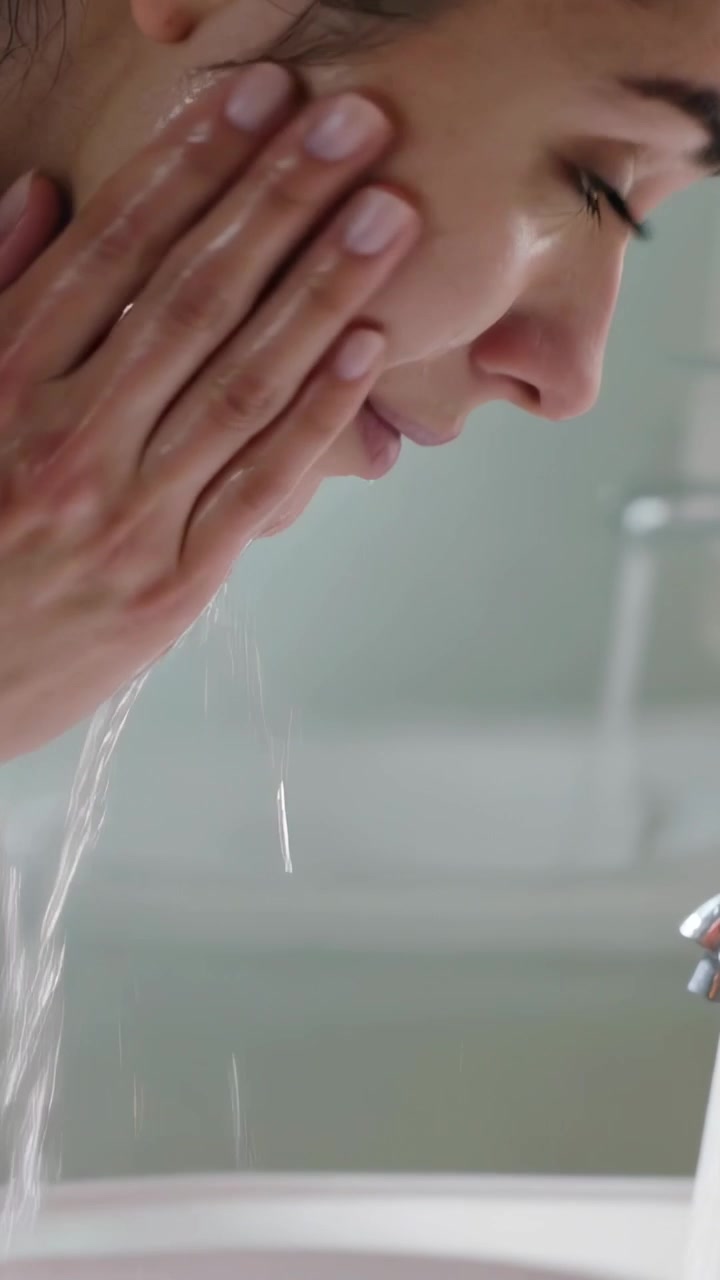 Vertical video: Rinsing woman cupping water under running faucet at bathroom sink, reaching faucet