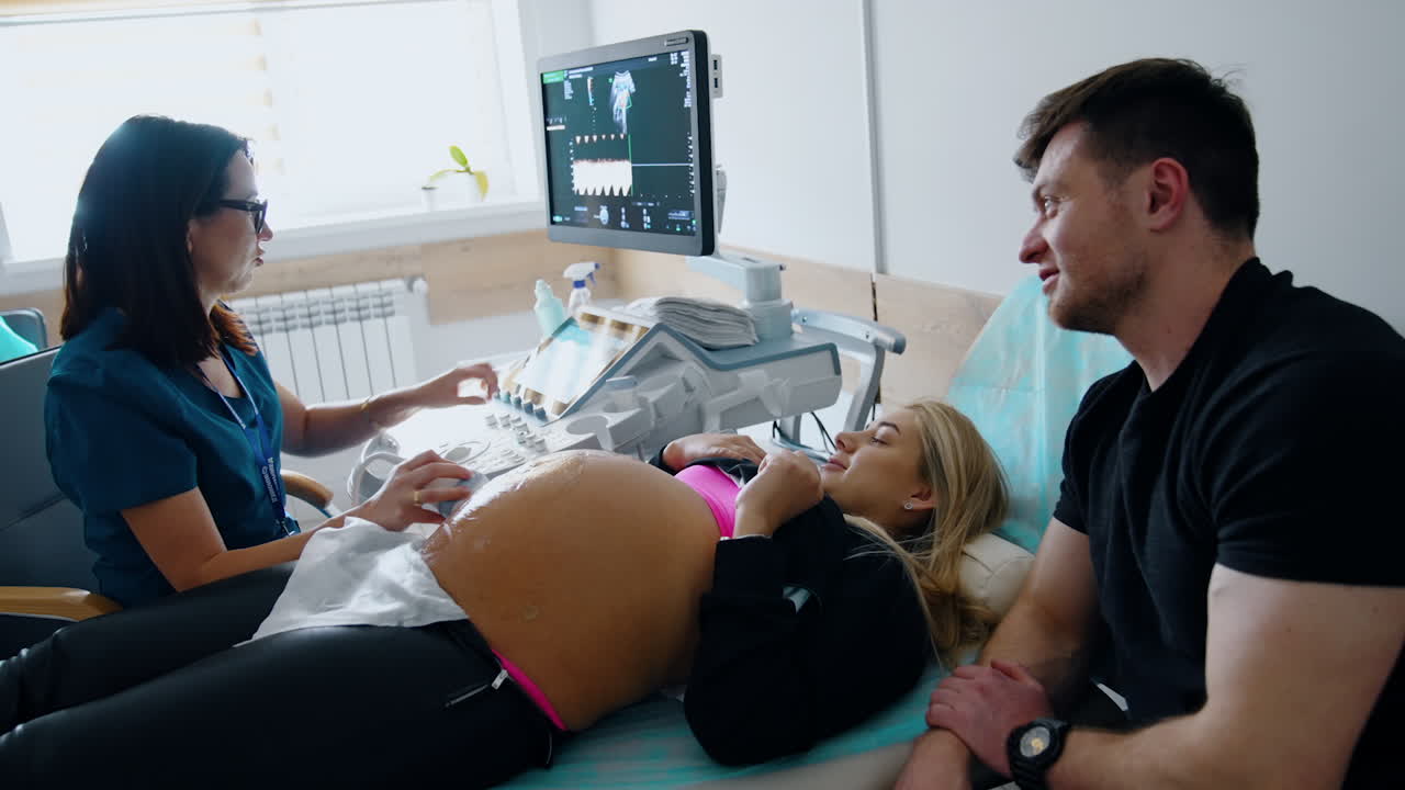 Expecting couple is at the cabinet of ultrasound examination. Pregnant woman lies on the couch checked by the doctor and husband sits beside.
