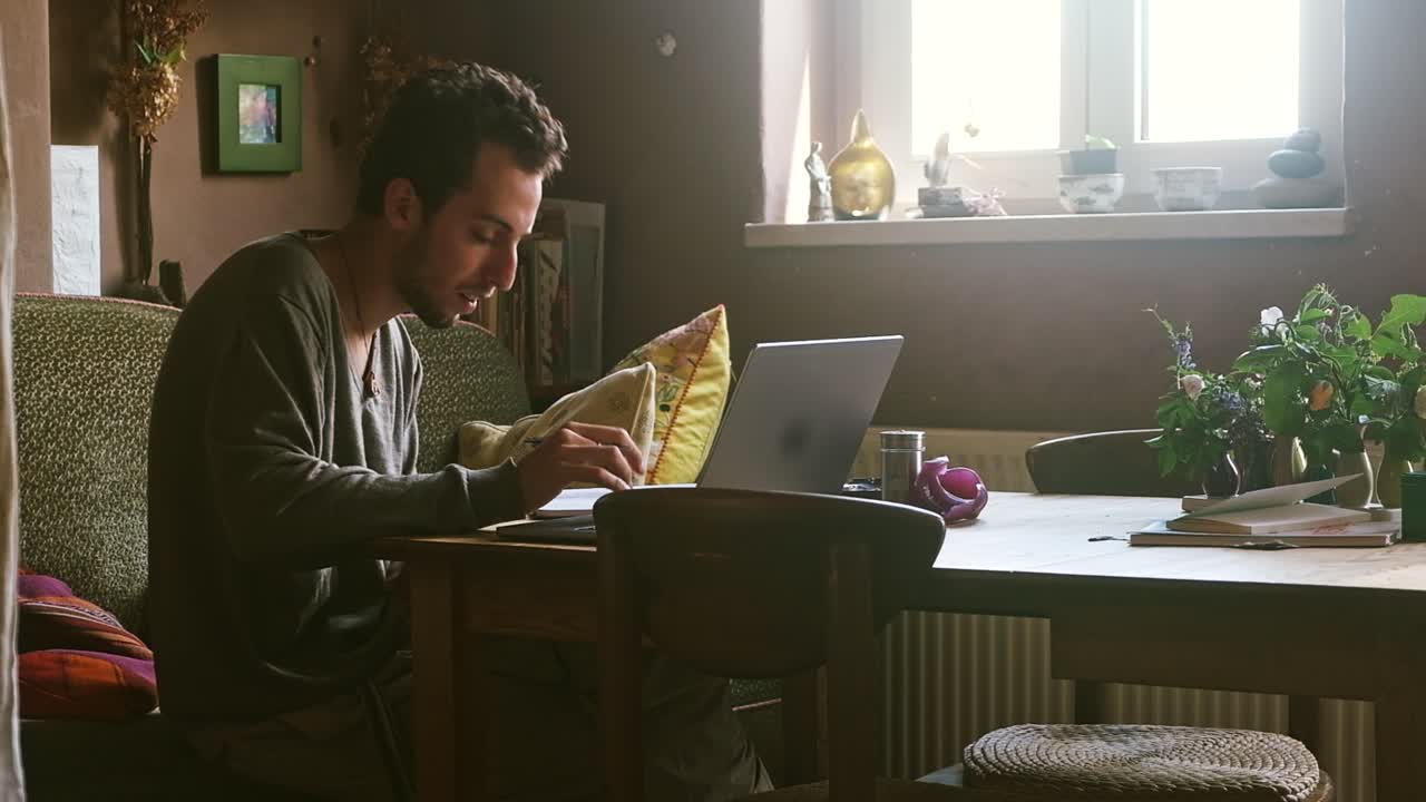 Man sitting at a desk, working on a laptop with plants and books around