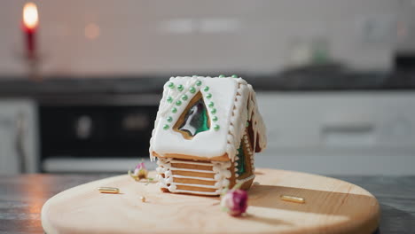 casa de pan de jengibre bellamente decorada con hielo blanco y cuentas verdes se sienta en una bandeja de madera, ambiente de cocina cálido con luz de vela suave y fondo borroso