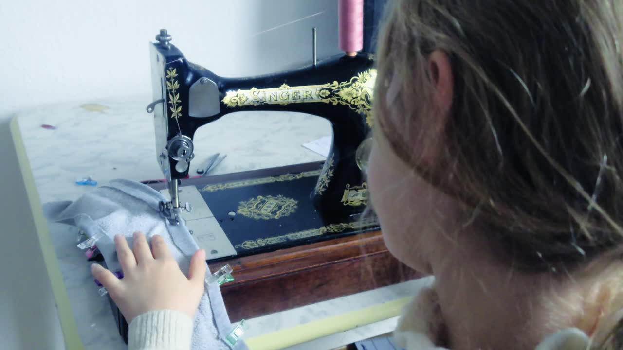 Inquadratura statica di un bambino seduto a una bellissima macchina da cucire manuale antica nera e oro, vecchia di oltre 100 anni, che impara la sartoria tradizionale a Budapest, Ungheria, in una giornata luminosa del 2026.
