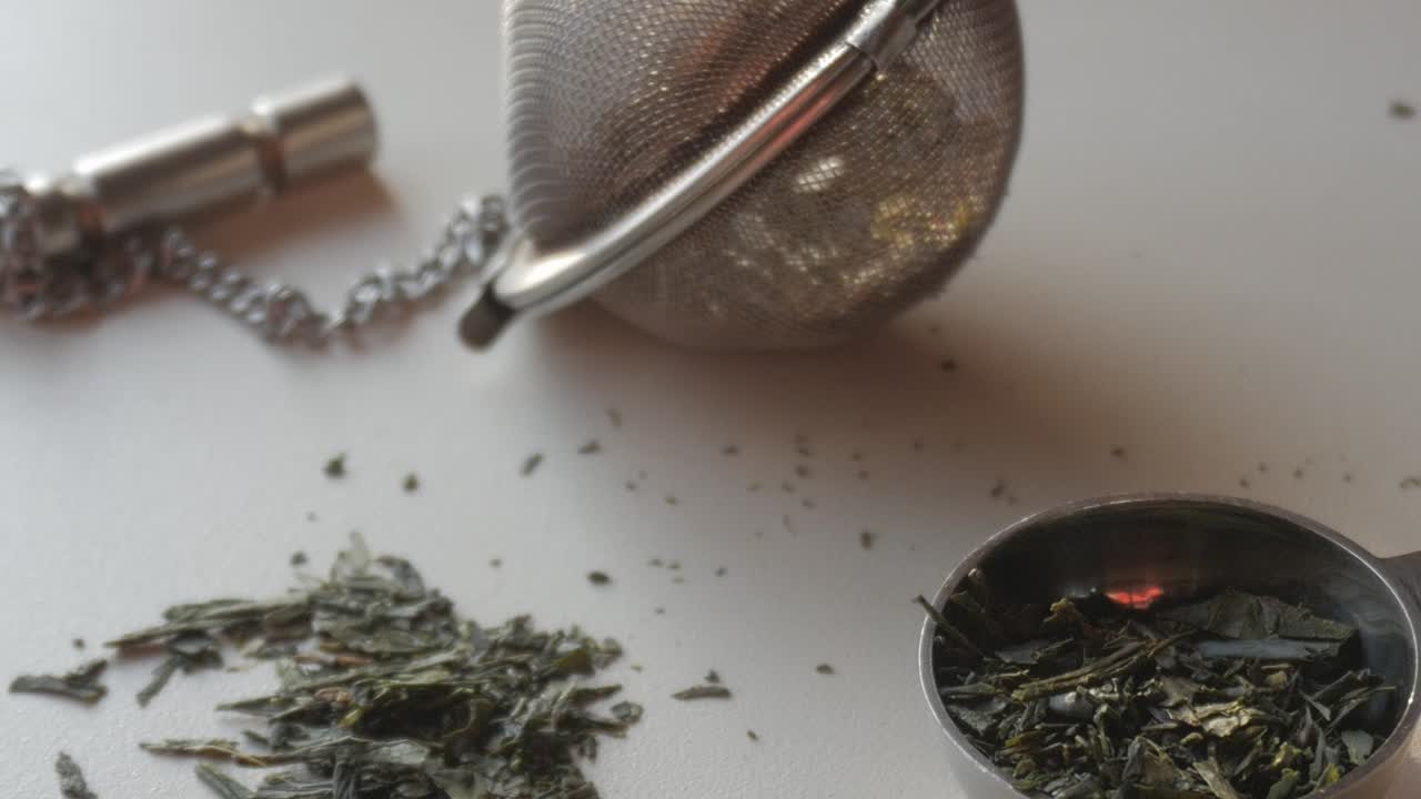 Loose green tea leaves scattered on a surface, next to a tea infuser and measuring spoon