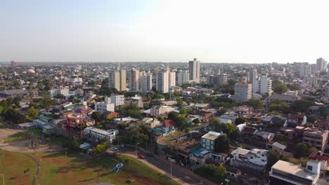 Aerial-drone-footage-of-large-city-skyline-near-a-park-at-sunset-in-Posadas,-Misiones,-Argentina