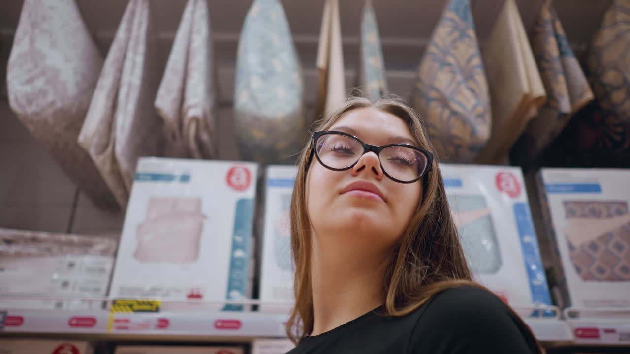 primer plano de una mujer caminando por un estante de la tienda lleno de productos de cama, mira a su alrededor, observando las opciones de edredones y almohadas, el entorno de la tienda con decoración para el hogar, textiles y artículos esenciales para el interior en exhibición