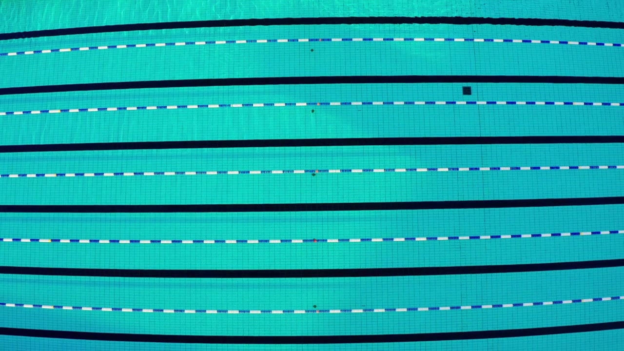 Uma vista aérea de cima para baixo de uma piscina vazia. A câmera se move lentamente sobre a água turquesa, mostrando as linhas pretas das raias no fundo azulejado e as cordas flutuantes das raias.
