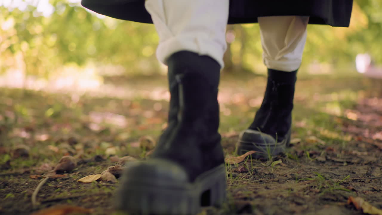 Leg view of stylish pedestrian in black boots and white pants with black long coat strolling on leaf carpet along shaded path, ground covered with fallen leaves, calm autumn park mood, nature walk