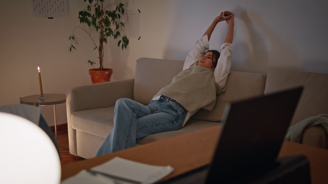 mujer descansando estirando el cuerpo en el sofá de la noche sola. mujer relajada sosteniendo el teléfono