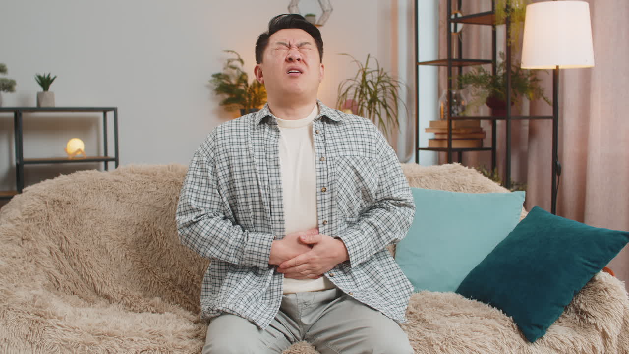 若い病気の男性がソファーに座って、家でソファーに突然の強い腹痛を感じている。