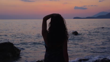 Silueta-De-Una-Chica-Con-El-Pelo-Rizado-Parada-En-Una-Playa-Rocosa-Viendo-Una-Hermosa-Puesta-De-Sol-Frente-Al-Mar