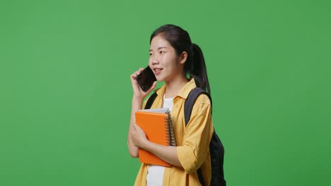 side view of asian woman student with a backpack talking on smartphone while standing in the green screen background studio