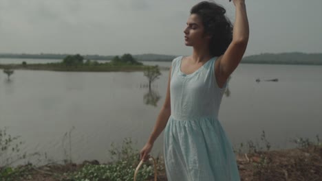 Toma-Estática-En-Cámara-Lenta-De-Una-Mujer-India-Elegante-Y-Elegante-Con-Vestido-Turquesa-Acariciando-Su-Cabello-Negro-Durante-El-Clima-Ventoso-Frente-A-Un-Lago-Con-Islas-Marinas-En-100-Fps