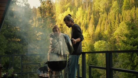 A-brunette-guy-and-a-blonde-girl-are-watching-the-preparation-of-meat-on-the-grill-during-a-picnic-on-the-terrace-against-the-backdrop-of-a-coniferous-forest-and-mountains