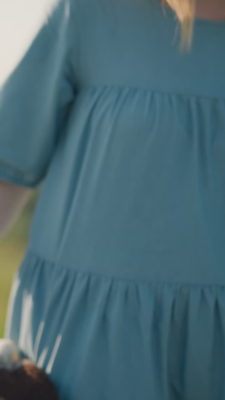A close-up shot of a mother in a blue dress bending down to pick up a basket filled with fresh fruit from the grass. The scene captures a simple yet warm moment of a picnic in a grassy field