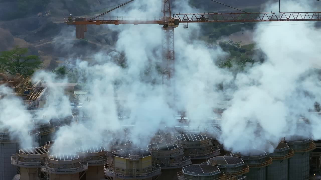 Cylindrical cooling towers release heavy white steam beneath large construction crane at Barrick Gold’s gold and silver mining complex near Cotuí, Dominican Republic. Slow pan left aerial