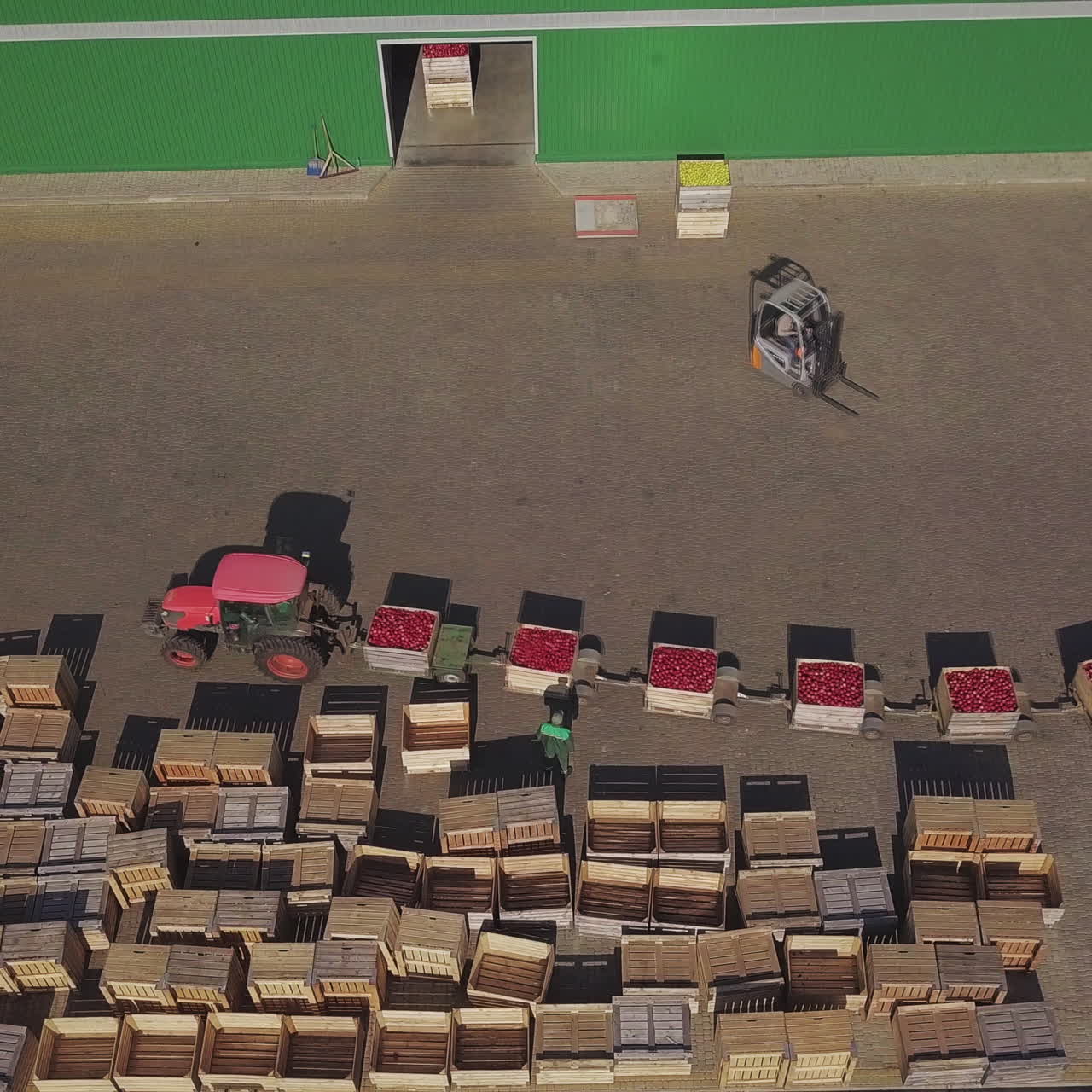 Red tractor with trailers full of red apples at farmland going near empty wooden crates. Farmhouse, tractor with trailers and a lot of crates outdoors. Aerial view