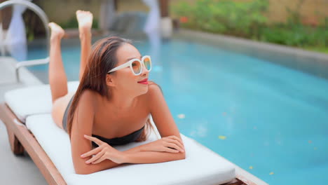 Hot-Asian-woman-in-a-black-bathing-suit-lying-on-her-belly-on-a-comfortable-deck-chair-in-a-gorgeous-tropical-resort-near-swimming-pool-smiling,-daytime-slow-motion