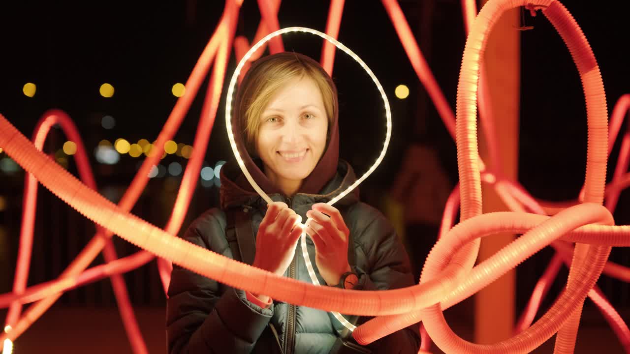 портрет красивой улыбающейся блондинки в капюшоне на фоне светящейся инсталляции. естественная красота