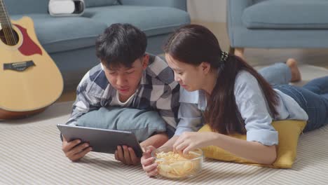 asian teen couple using tablet while lying on carpet on the floor at home. eating snacks watching movie, speaking, and enjoying time together