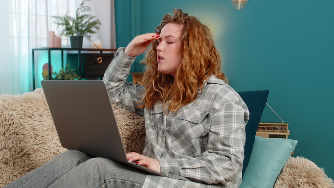 Young woman sits on sofa with laptop upset holding hand on forehead feeling failure and frustration