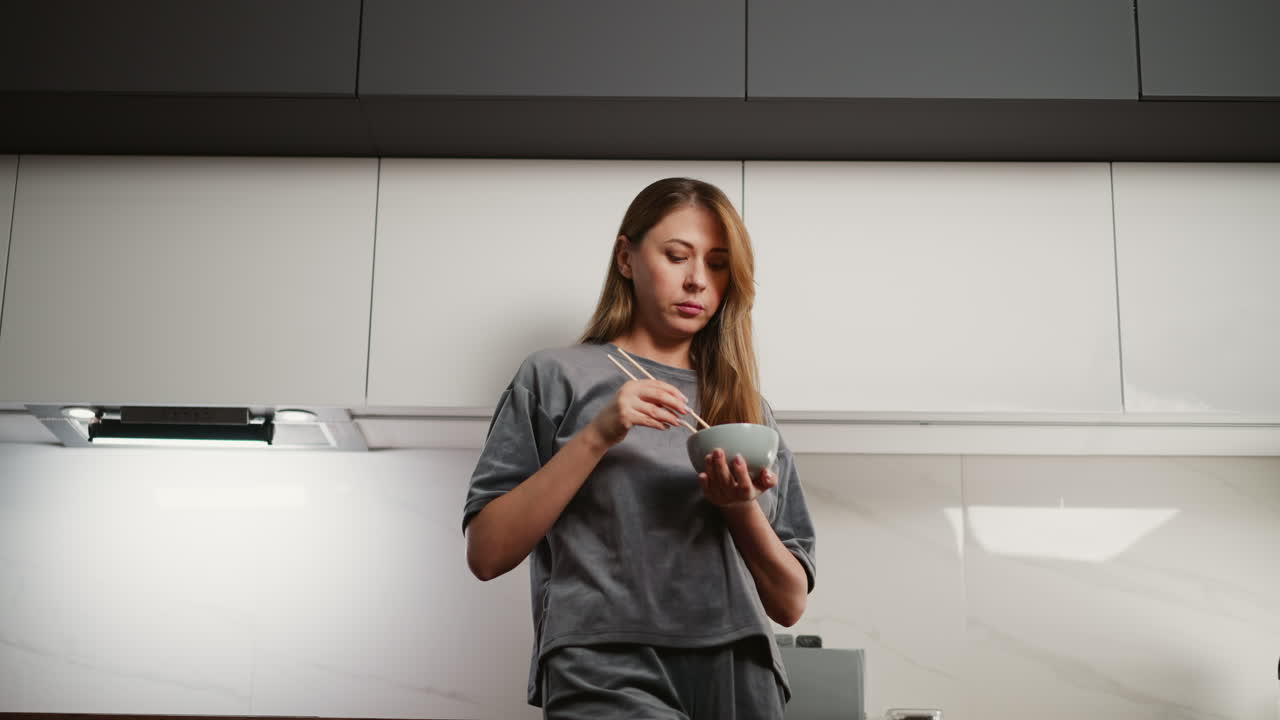 Woman with long flowing hair stands in modern kitchen eating noodles with chopsticks, dressed casually and enjoying quiet moment while savoring meal