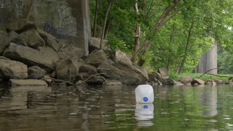 milk-jug-slowly-floats-down-river-towards-rocky-bridge-base-slow-motion