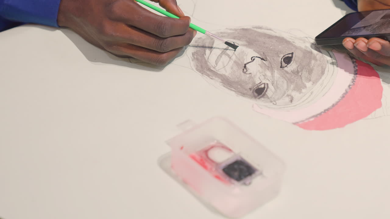 Svart konstnär från ovan lägger till ansiktsdetaljer, grönt verktyg förfinar skuggning på ett monokromt porträtt, smartphone som referens hålls vid kanten, liten palettlåda och rosa hatt som accent, koncentrerat och noggrant.