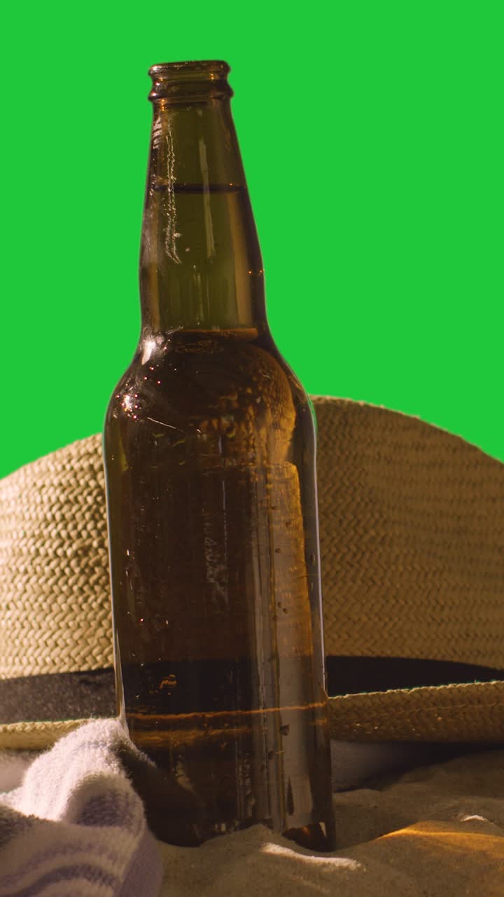 video concepto de vacaciones de verano vertical de botella de cerveza en la toalla de playa con y sombrero de sol contra pantalla verde
