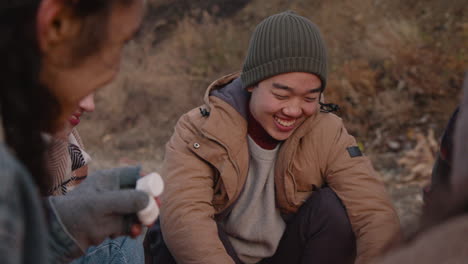 camera focuses on an teenage boy talking to a friend who is sitting in front of her near the bonfire