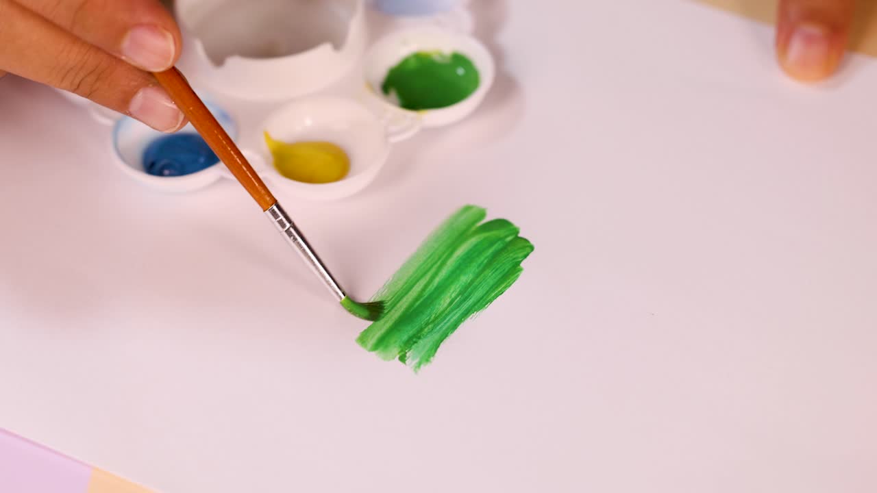 Artist uses brush to blend green watercolor on paper, bright lighting, overhead camera angle