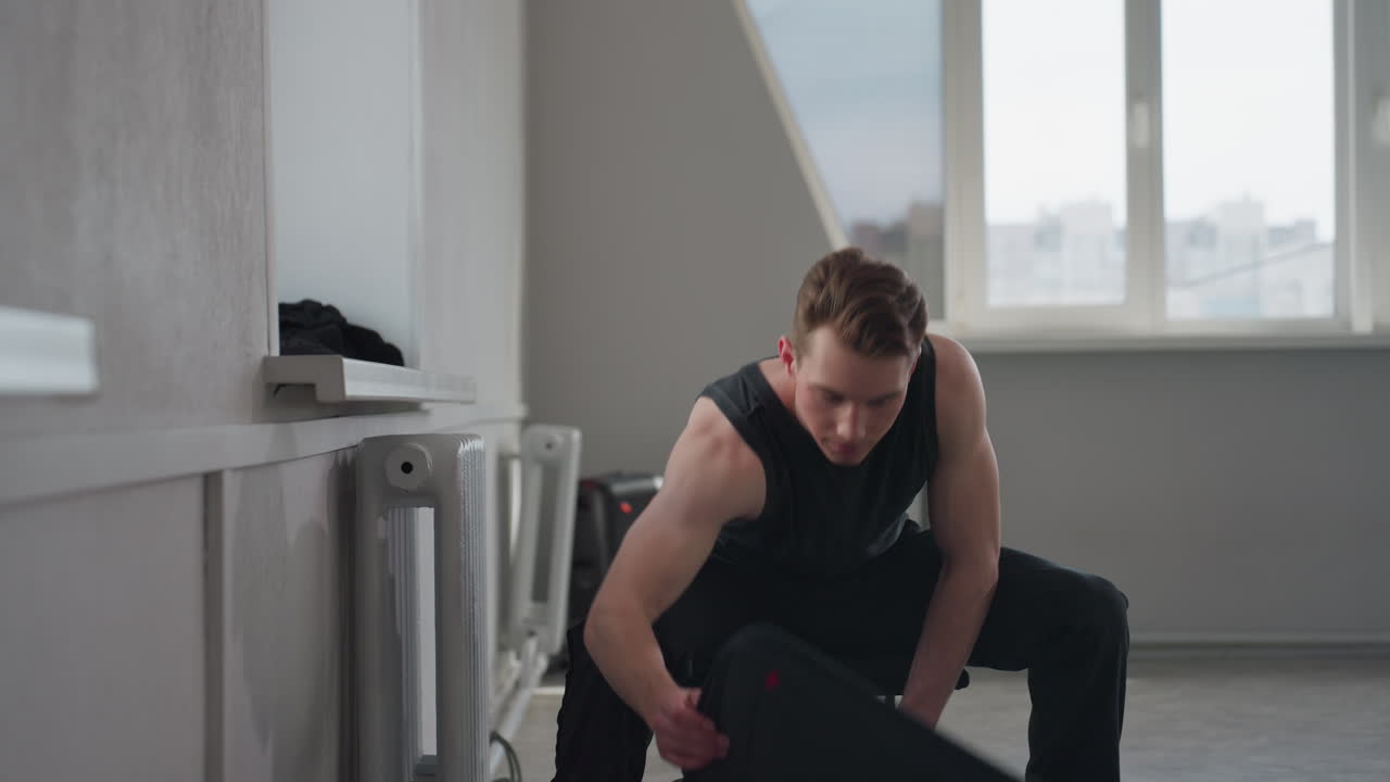 Young man in black sleeveless top sits on stool beside large window in empty bright room, reaches to place zipped garment bag and clothing on radiator ledge before standing up