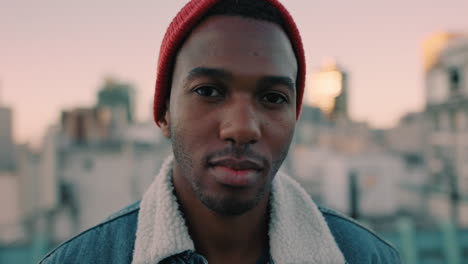 portrait attractive young african american man on rooftop at sunset wearing red beanie hat looking confident in urban city background