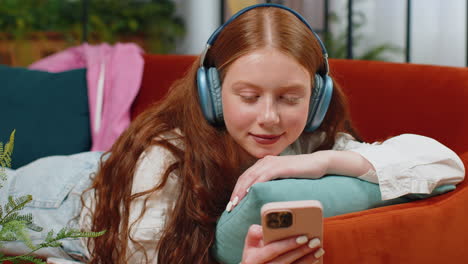 Happy-teenager-girl-in-wireless-headphones-relaxing-lying-on-sofa-at-home-listening-favorite-music