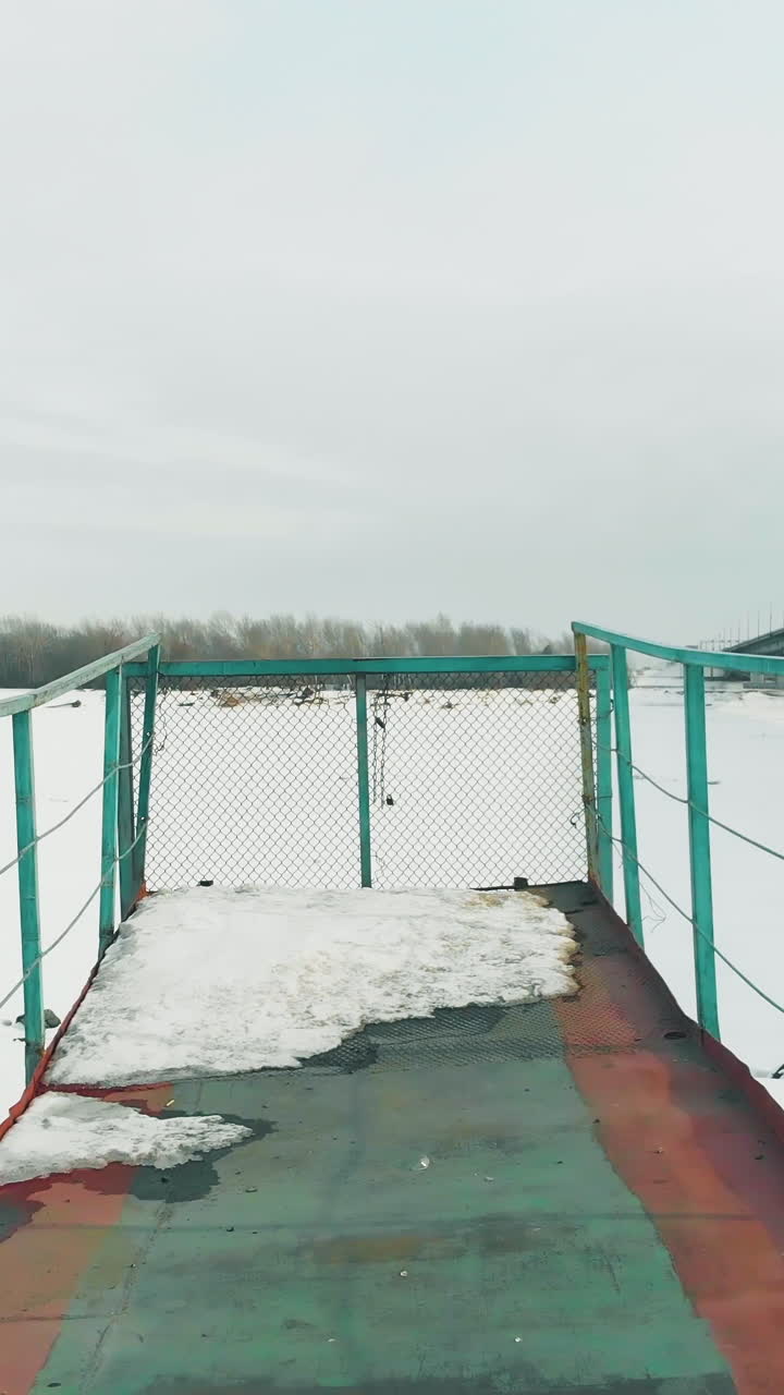 rusty pier with green rails stands at frozen river against large bridge under grey winter sky upper view