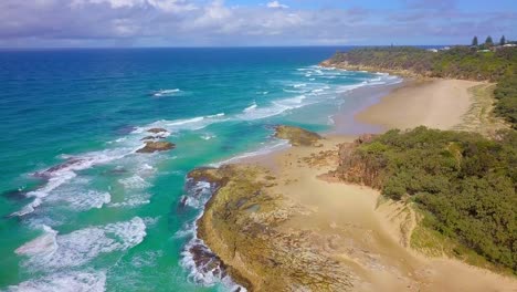 aerial fly over a wild, sandy beach with cliffs, rocks, turquoise blue water and splashing waves