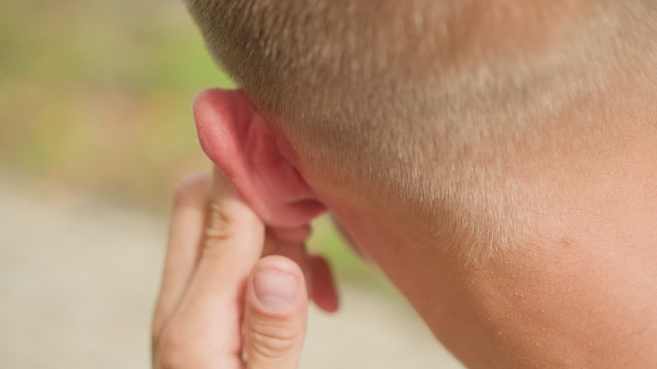Primer plano de un hombre ajustándose el audífono mientras camina por la acera, con la sien sudada y la montura de las gafas visibles. La yema del dedo presiona el suave audífono en el oído irritado, con un movimiento correctivo rápido sobre un parque borroso.