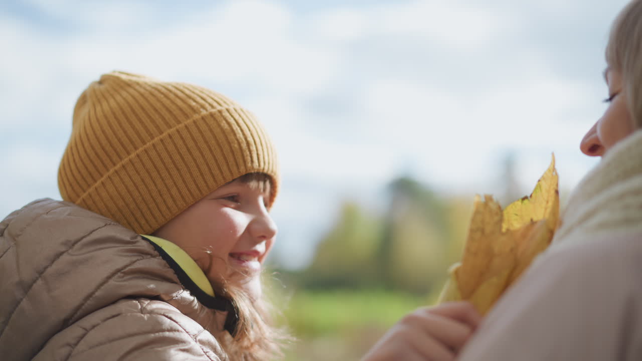 affectionate moment as child wearing mustard beanie gently embraces aunt holding autumn leaf under daylight sky, both smiling warmly while sharing emotional