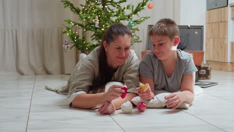 mother and son lying on marble floor smiling warmly while playing with christmas ornaments near decorated christmas tree cozy indoor setting festive family bonding joyful holiday atmosphere