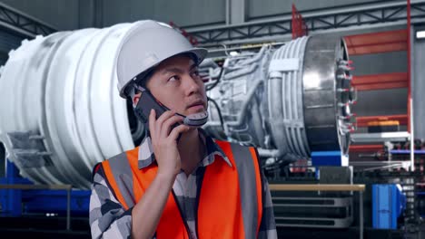 close up of asian male engineer with safety helmet talking on smartphone while standing with airplane engine maintenance conducted, analytics checking the turbine