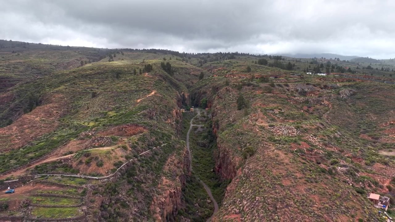 Vista aérea volando sobre un barranco profundo y colinas volcánicas con caminos rurales y terrazas.